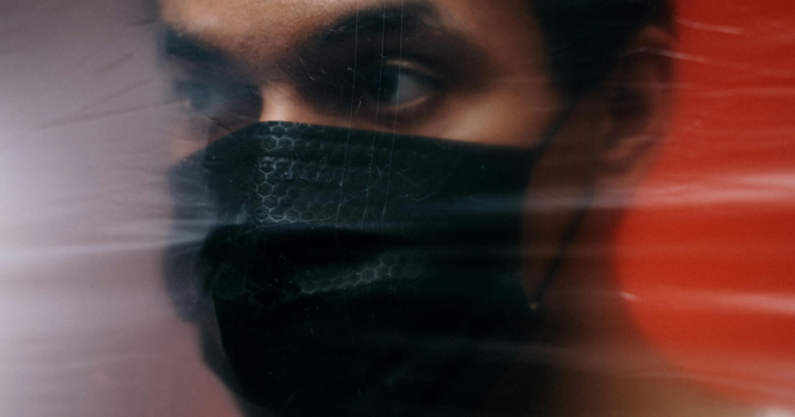 A man wearing a cloth mask with his masked face pushed up against plastic while looking grim