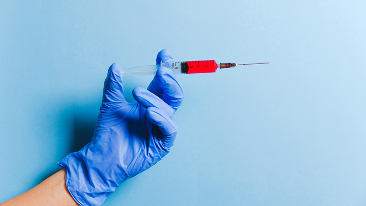 A gloved hand against a dull blue background holds a syringe with red fluid, preparing to inject it.