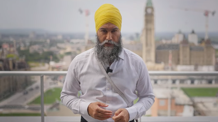 Jagmeet Singh speaks to the camera in a white collared shirt and an orange pagri on a balcony in front of Parliament hill